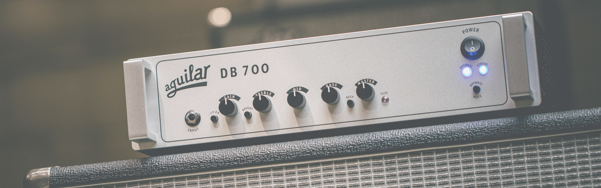 A close-up of an Aquilar DB 700 guitar amplifier head resting on top of a speaker cabinet, showing the front control panel with knobs and illuminated power lights.